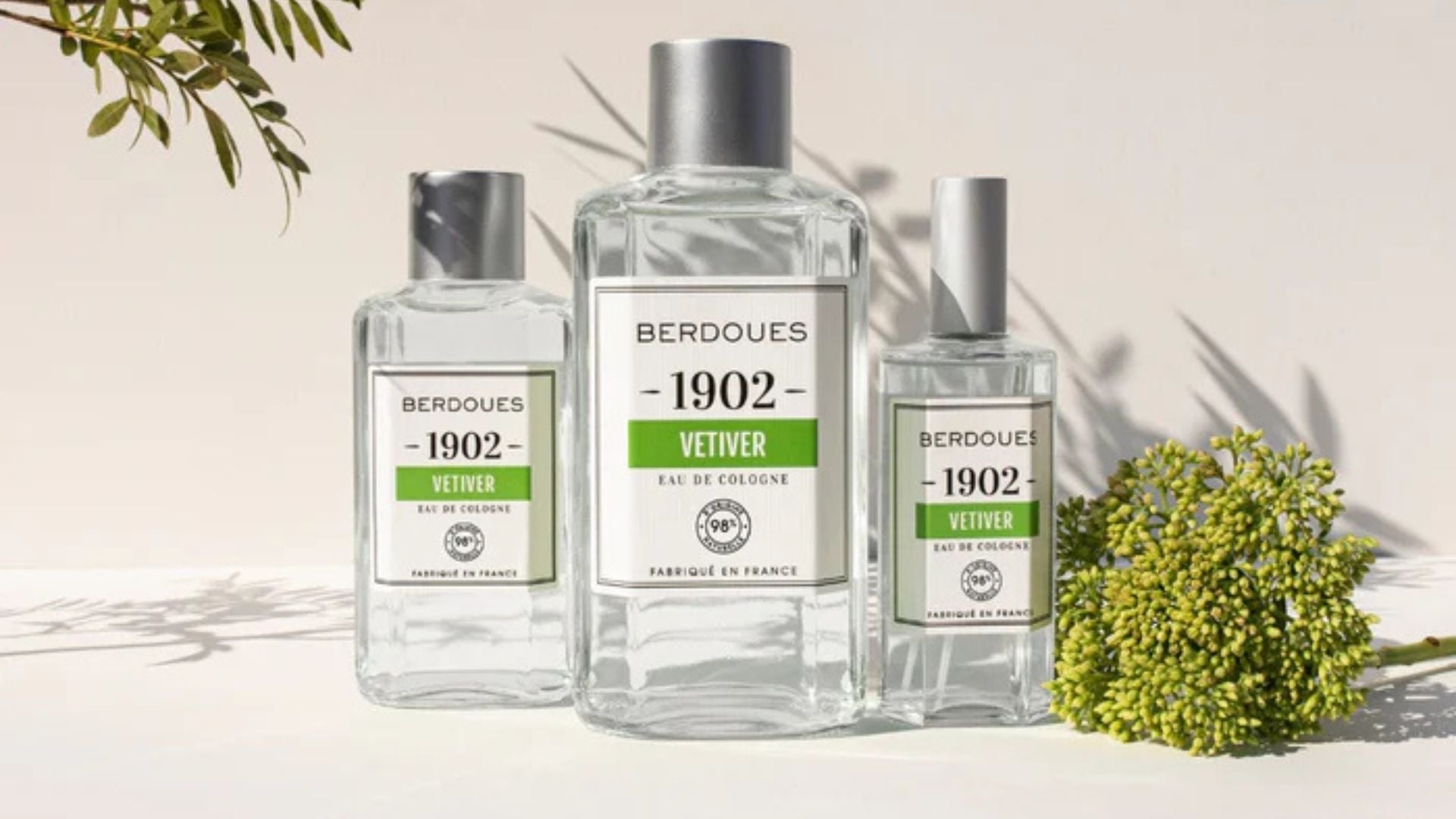 Trois flacons d’eau de cologne Berdoues 1902 au vétiver, posés sur une surface blanche avec une ombre végétale en arrière-plan et une branche fleurie en premier plan