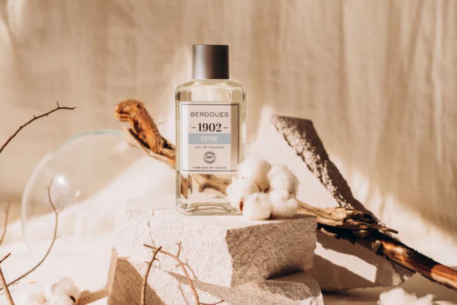 Flacon d’Eau de Cologne 1902 Coton Berdoues posé sur des pierres avec des fleurs de coton, symbole de douceur et de pureté.