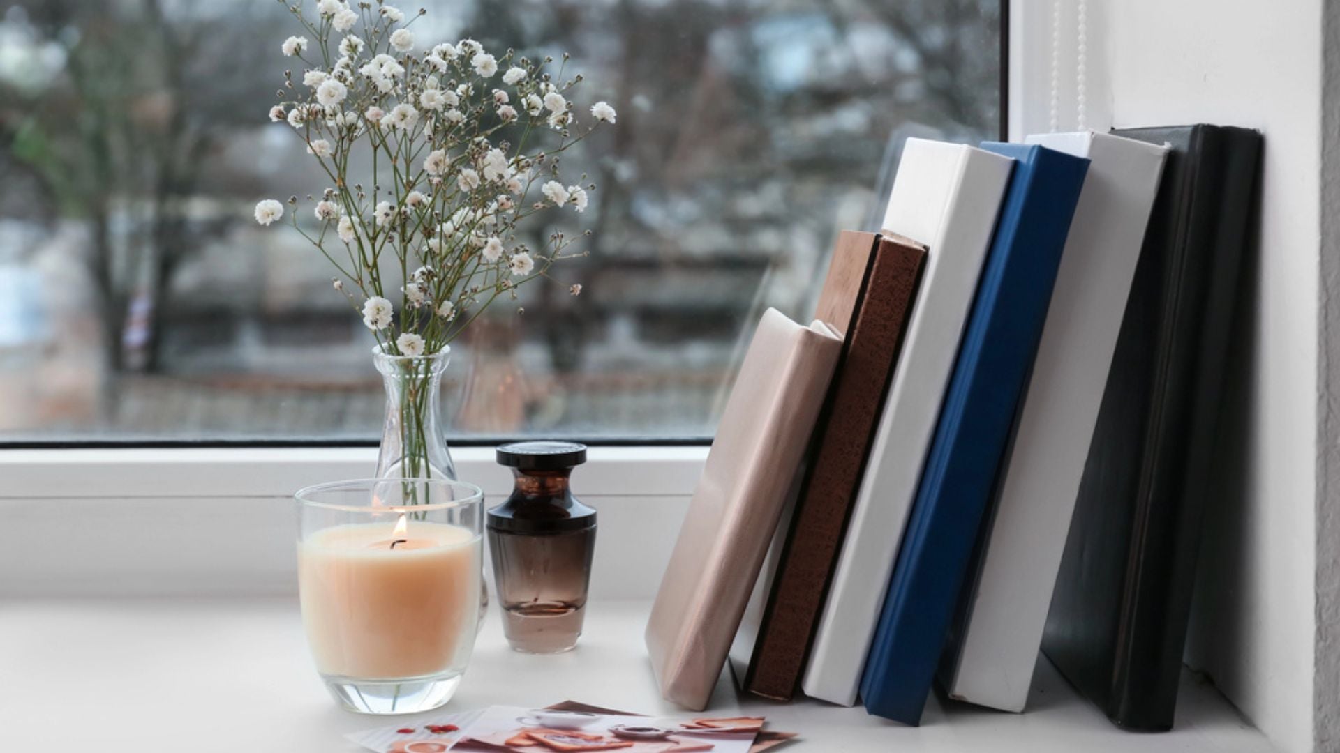 Bougie allumée, flacon de parfum, bouquet de fleurs et livres posés sur un rebord de fenêtre, évoquant l’univers raffiné et poétique du parfum à la française.